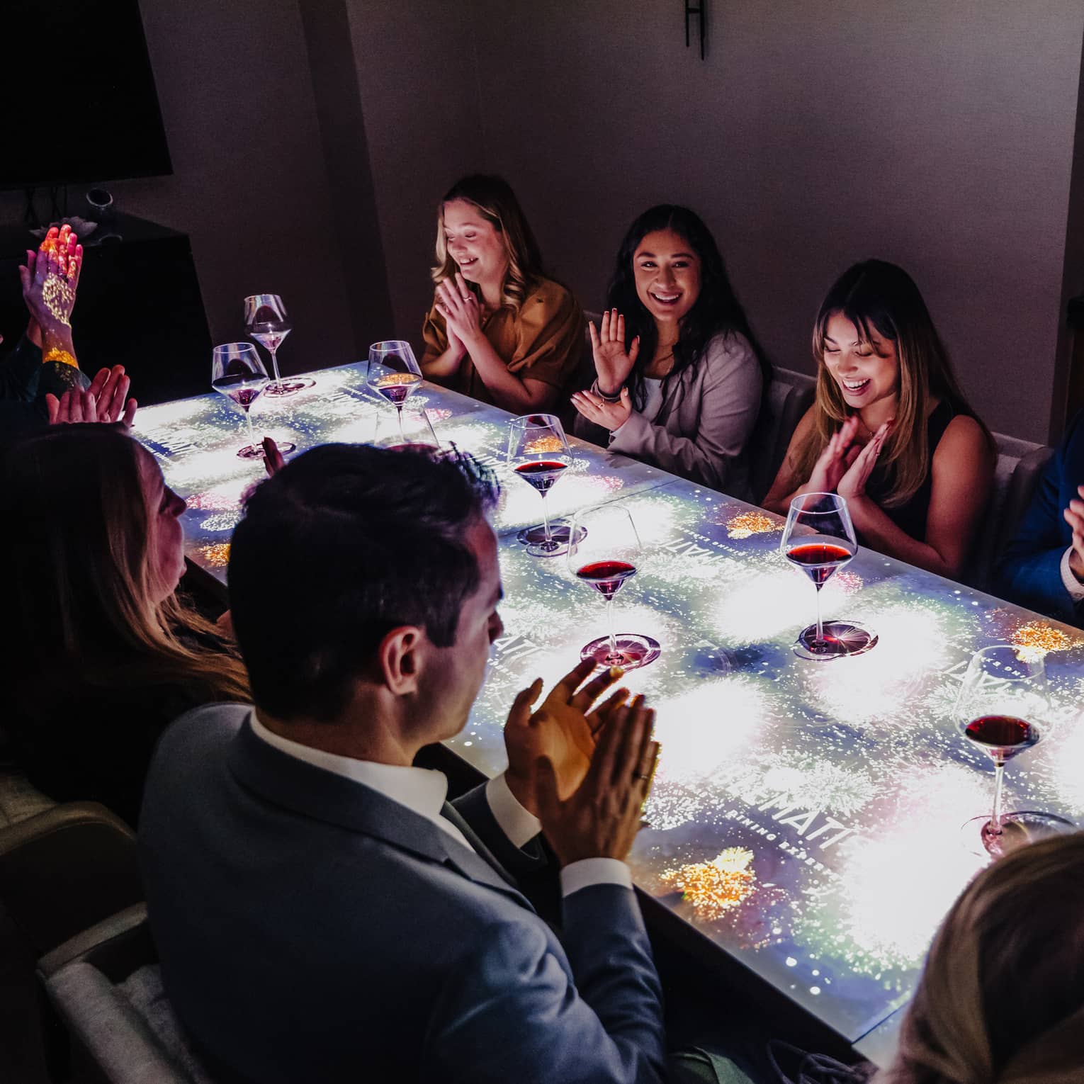 A large group of people sitting at a table with a screen on top of it drinking wine.