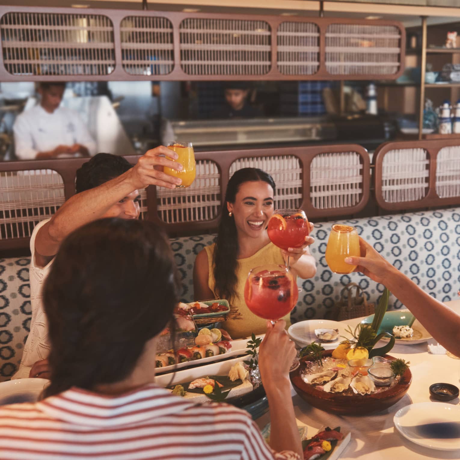 Four people sit in an blue-and-white upholstered restaurant booth, each raising a glass and smiling over a table filled with seafood platters