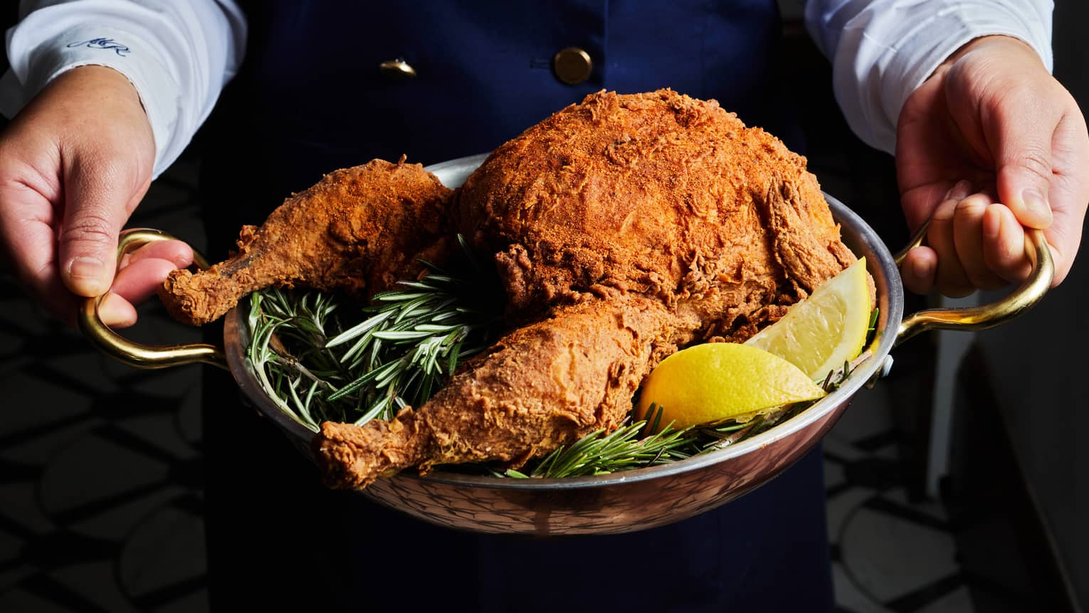 A person holds a serving tray of fried chicken with green garnishes and lemon slides.