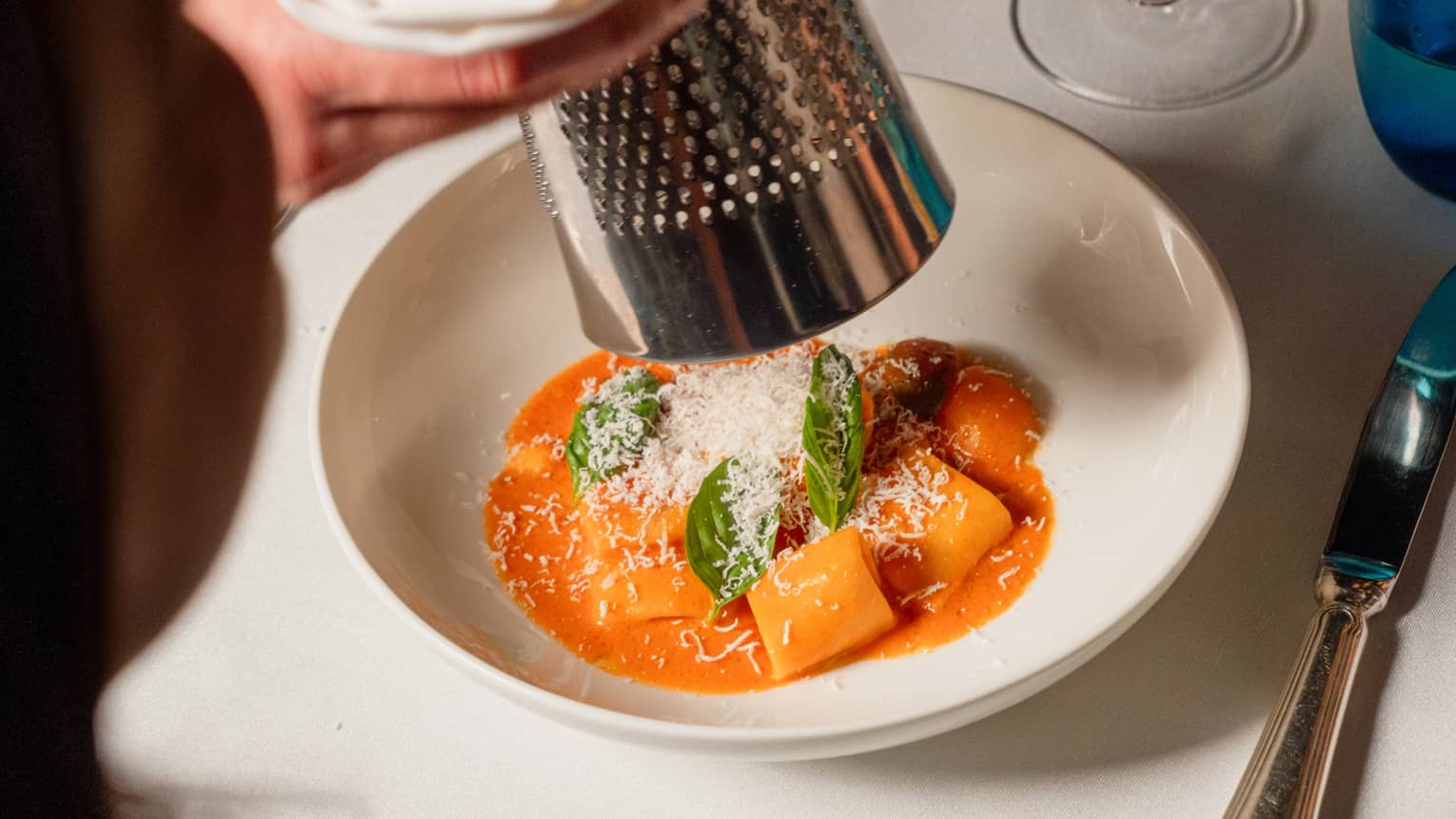 Person grating cheese over a pasta dish with a wine glass and silverware on the side