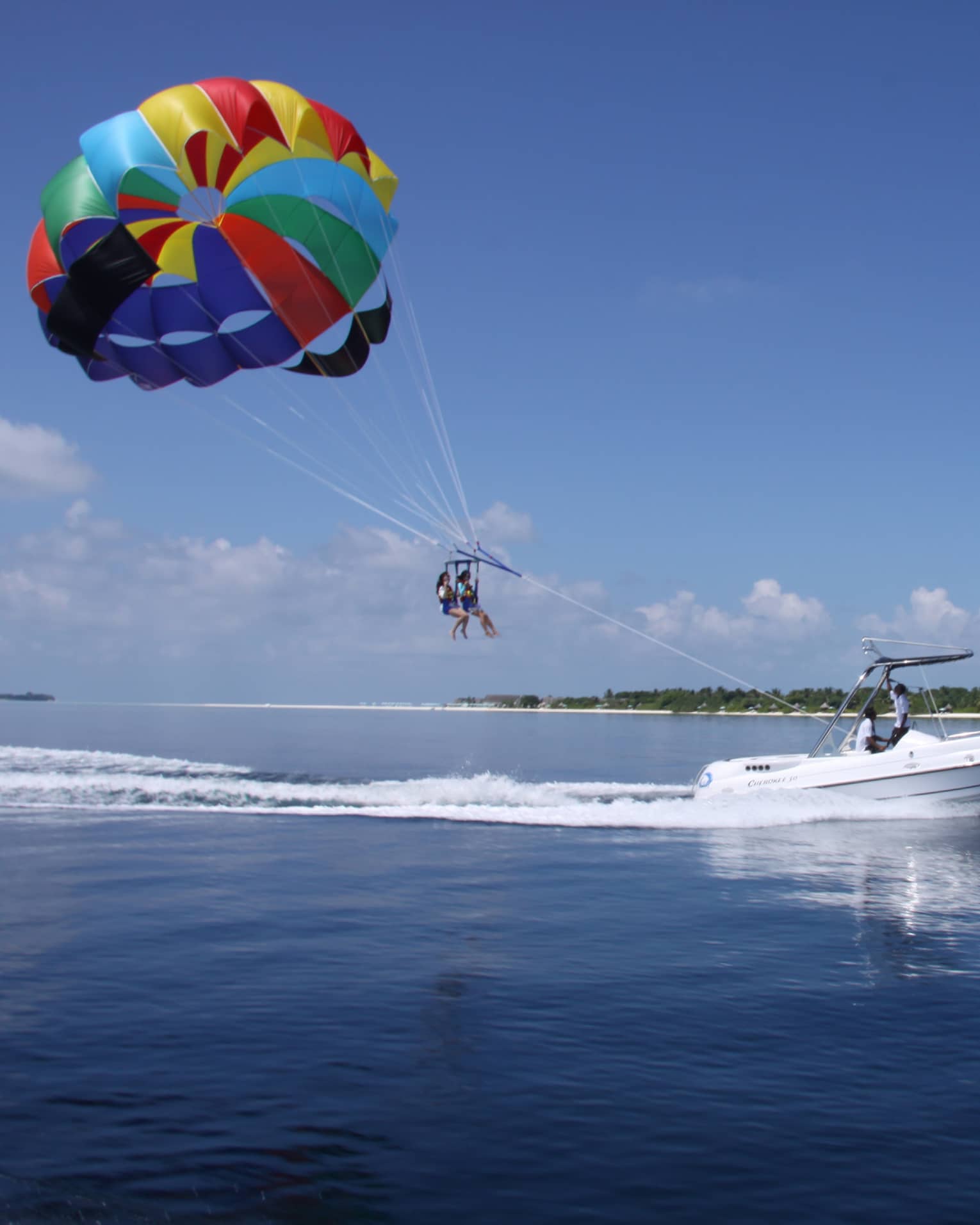 Guided by a motorboat trailing a frothy wake, two parasailers float through the air under a colourful parachute-style canopy.
