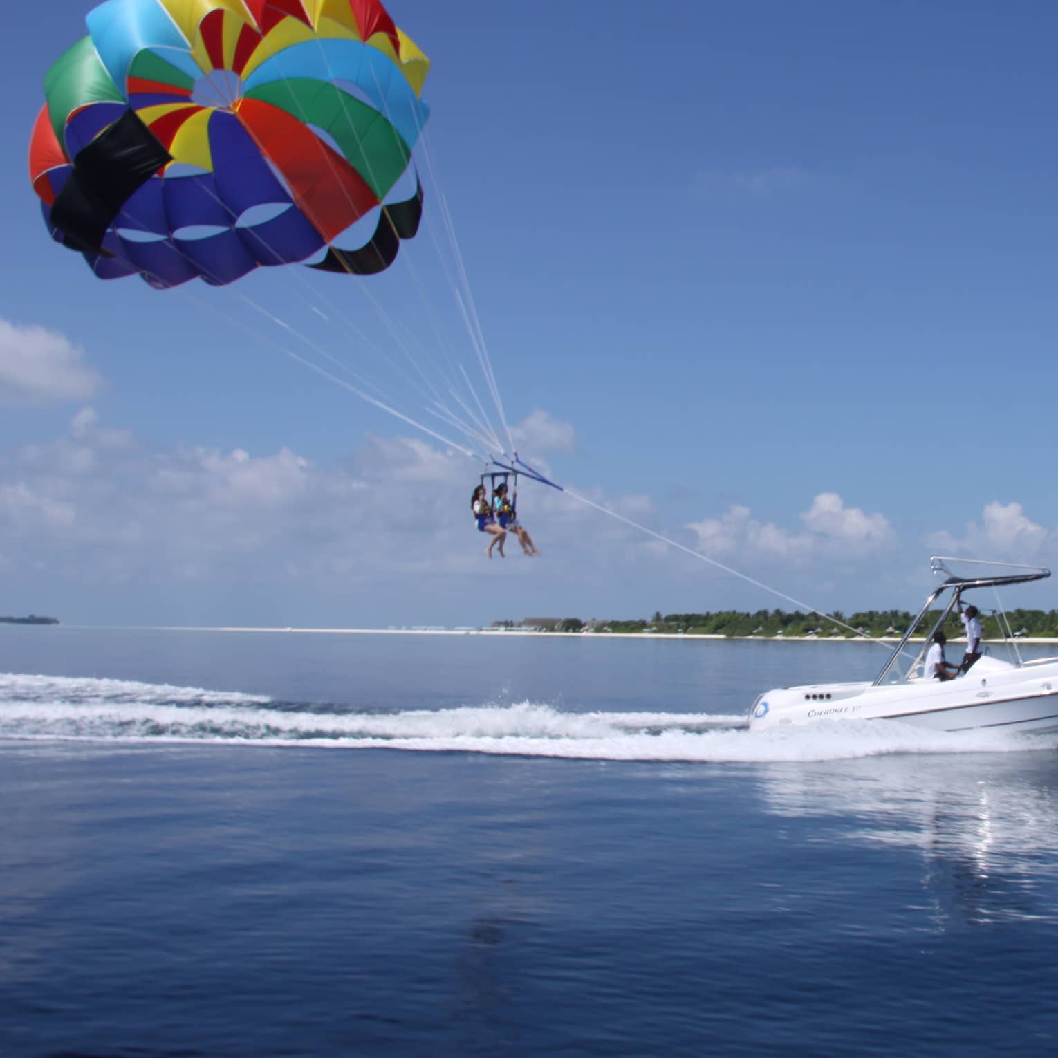 Guided by a motorboat trailing a frothy wake, two parasailers float through the air under a colourful parachute-style canopy.