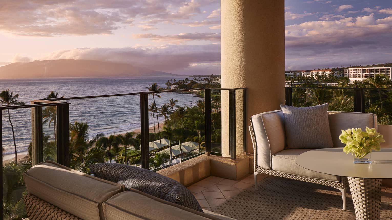 Suite balcony with cushioned sofas and a table overlooking the ocean and palm trees at sunset