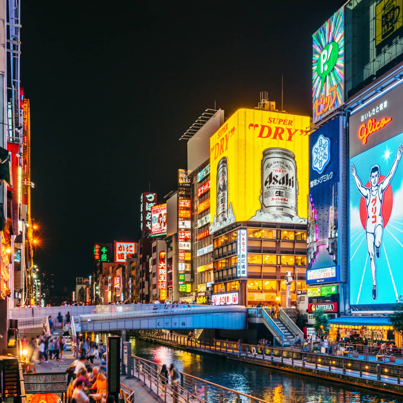 A footbridge crosses a narrow canal flanked by huge neon ads in bright colours on tall buildings in a busy city at night.