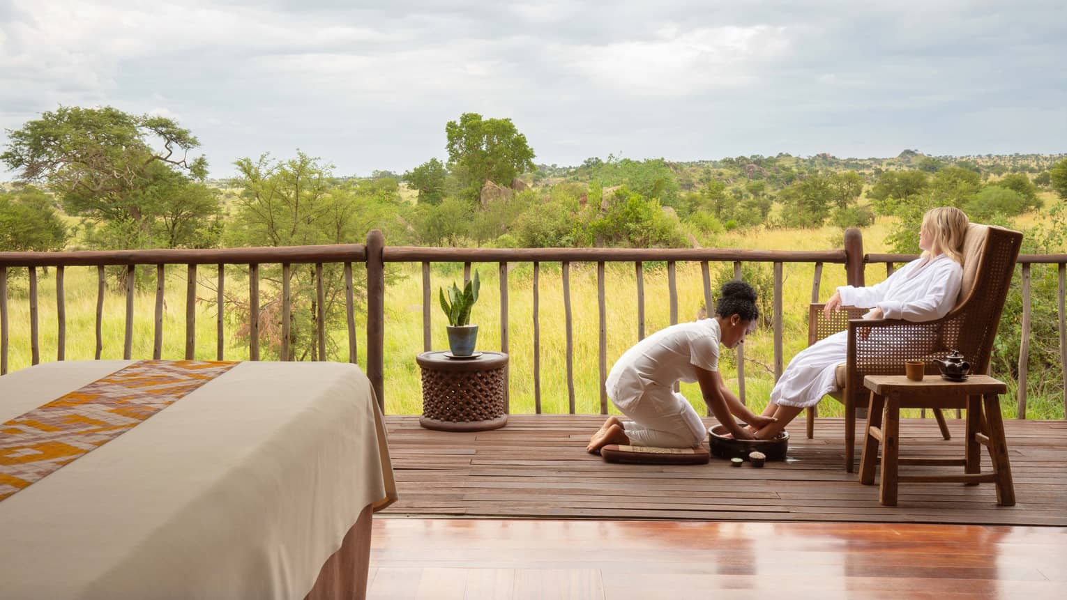 Spa therapist bathing fee of seated robed guest on spa terrace, looking out to expansive plain over vertical wooden railing