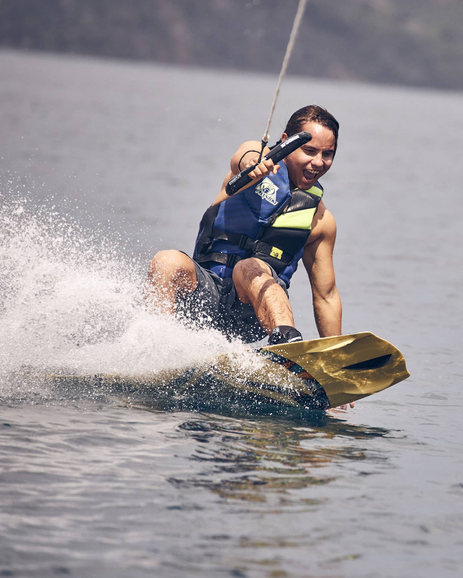 Young man wearing a blue and green life vest and blue swim trunks wakeboarding through the water