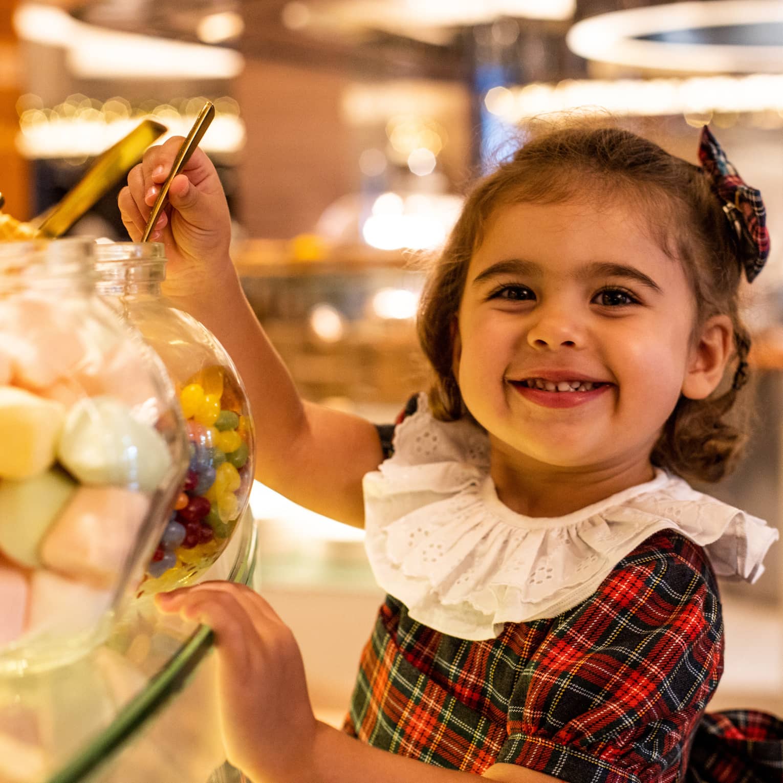 Little girl with chin-length brown hair and wearing a red-and-green plaid dress with a ruffled white collar smiles at the camera as she scoops up some candy from a jar
