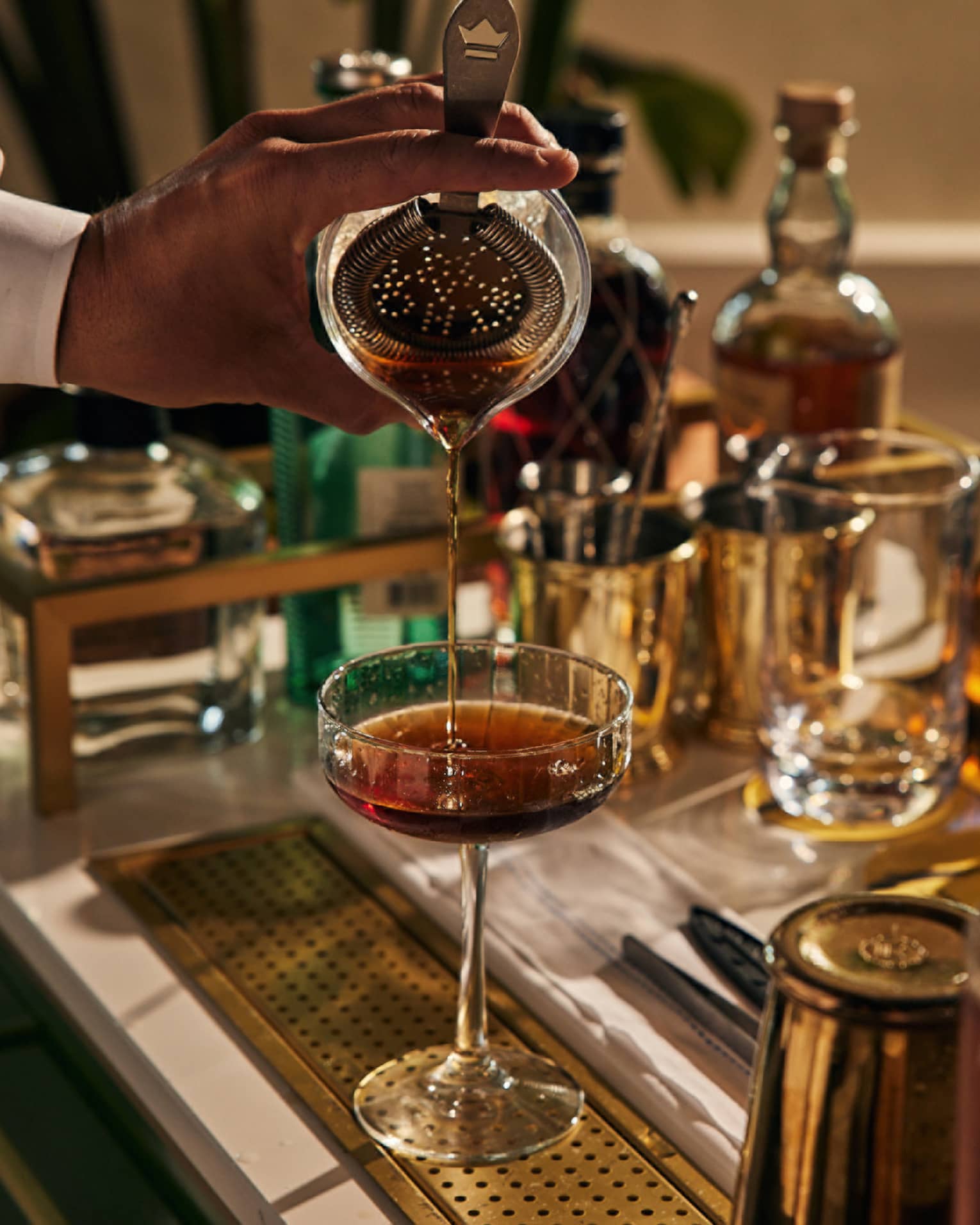 A bartender pouring a drink into a cocktail glass with bottles of liquor around.