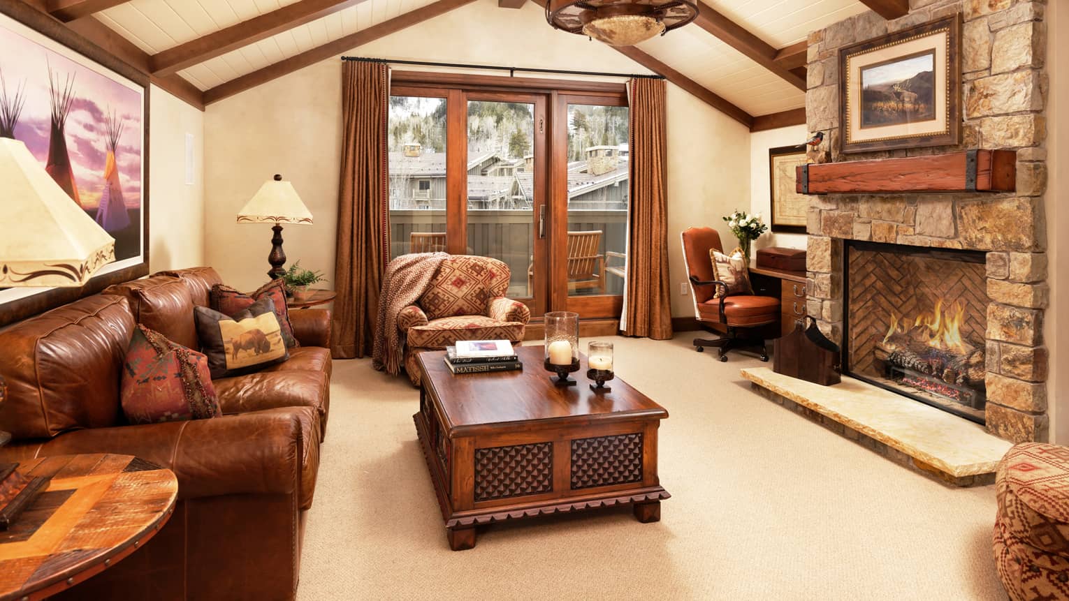 Rustic living room with leather sofa, armchair, tables, desk, stone fireplace, vaulted wood-beam ceiling and balcony doors with mountain view