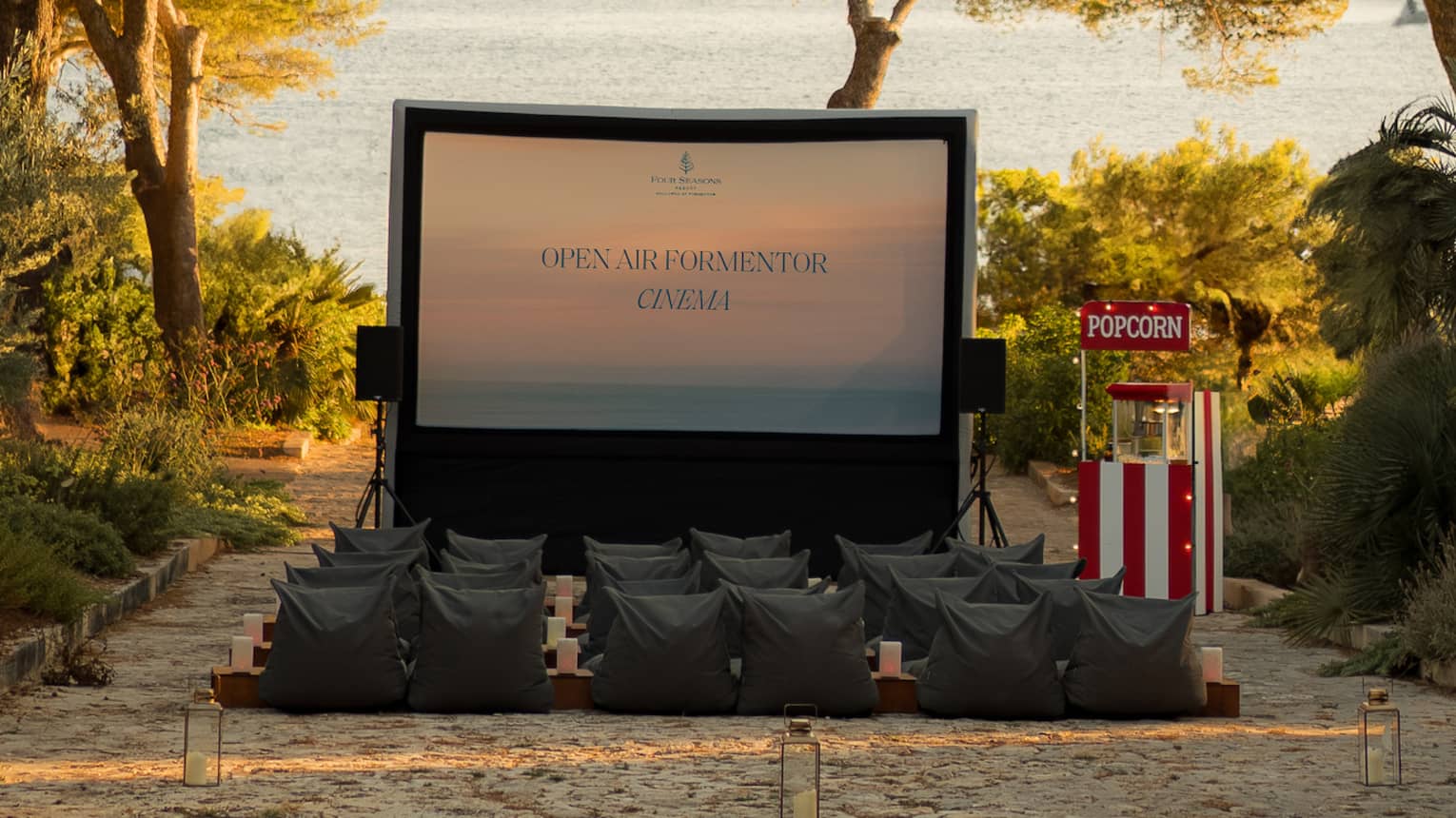 A large movie screen and rows of chairs in an outdoor setting  in the daytime. The movie area is surrounded by trees, and the ocean is in the background.