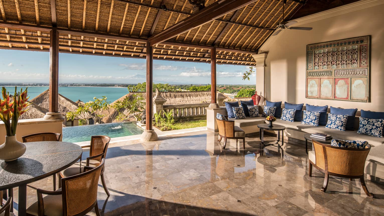 Gleaming tile floor under thatched roof of open-air space with large seating area, round dining table, infinity pool off front