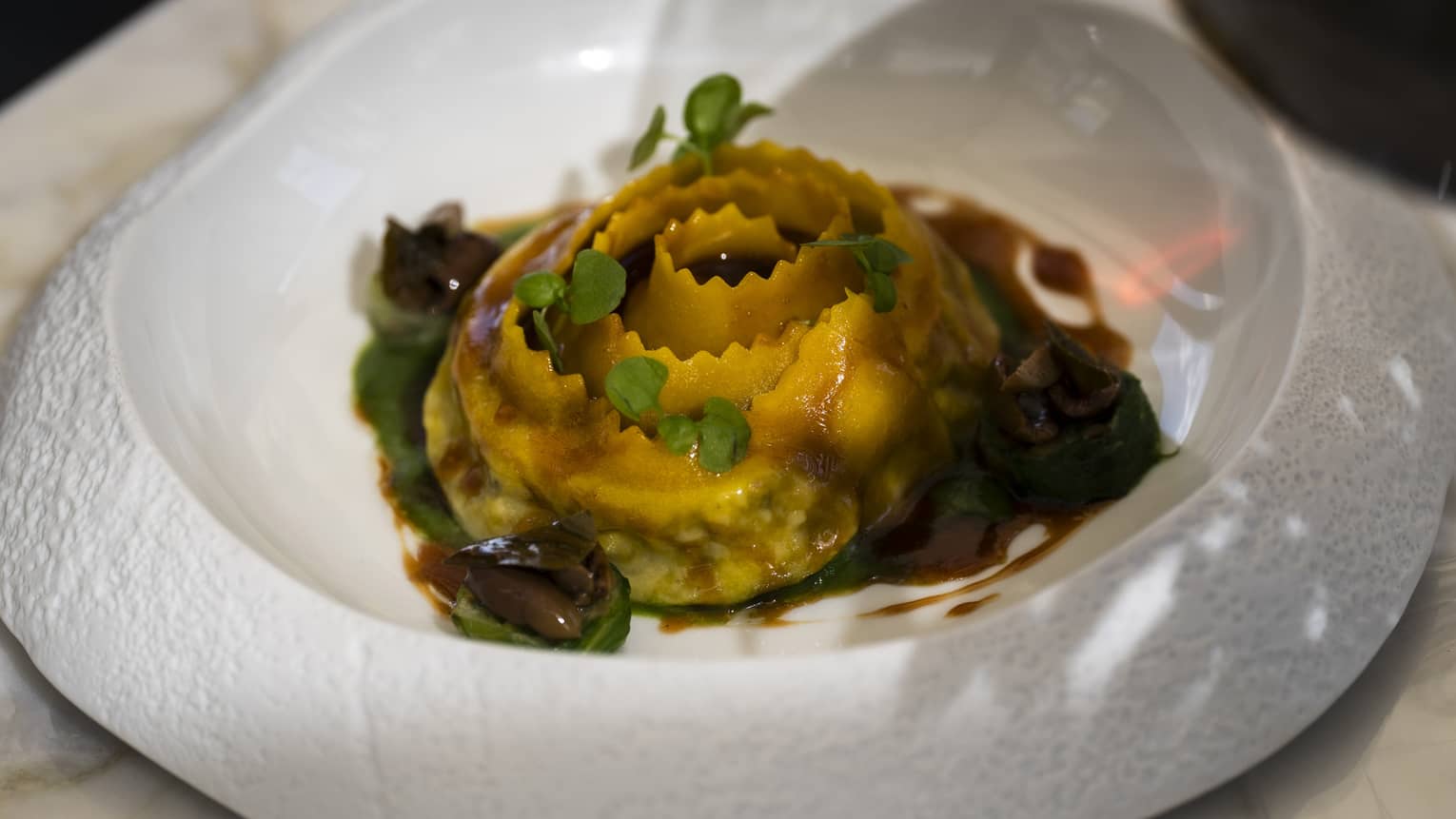 An artistic round white bowl with a textured rim holds a colourful pasta dish with garnishes and a dark sauce.