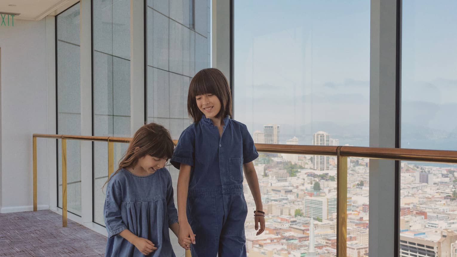 Two children walk hand-in-hand down a hallway in front of floor-to-ceiling windows overlooking San Francisco.