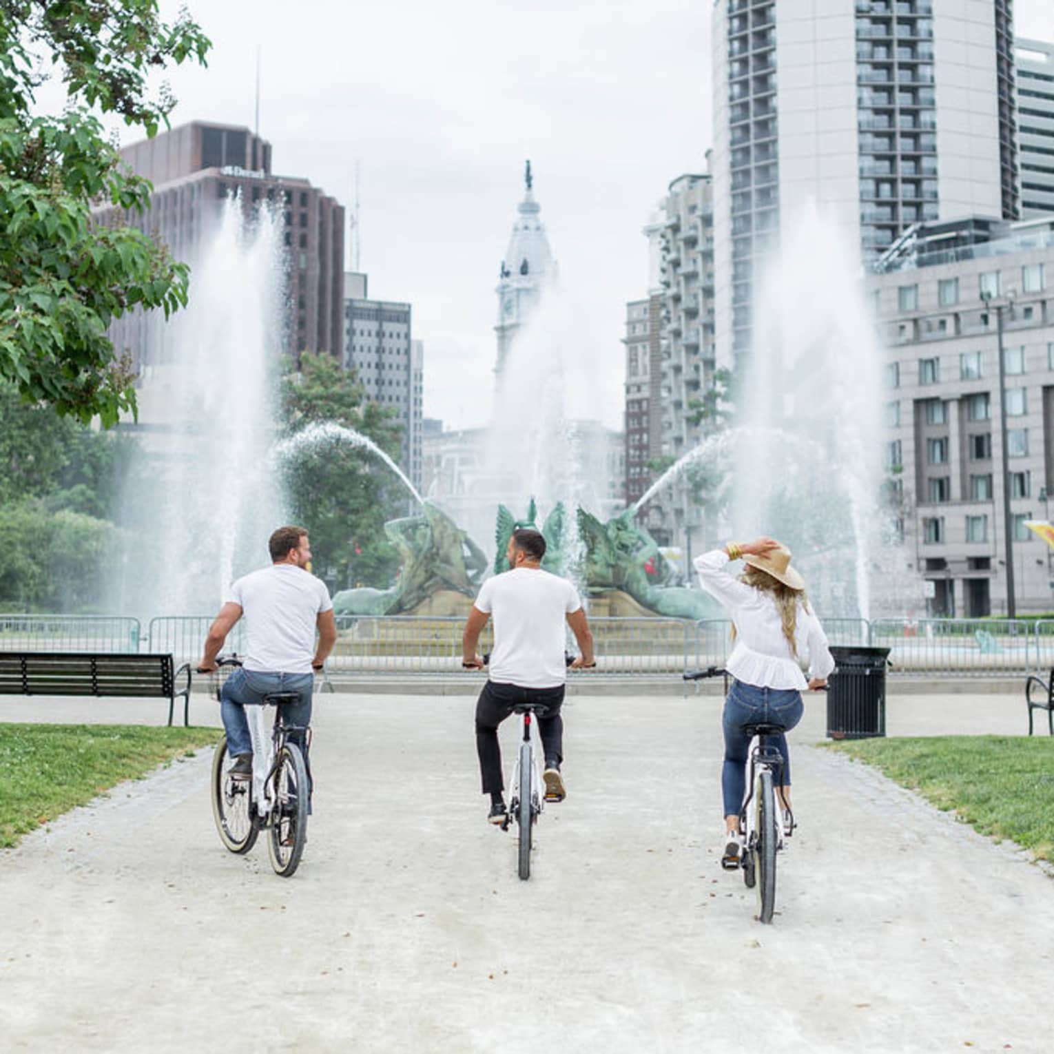 Three guests riding bikes towards a large fountain with the Philadelphia skyline in the distance