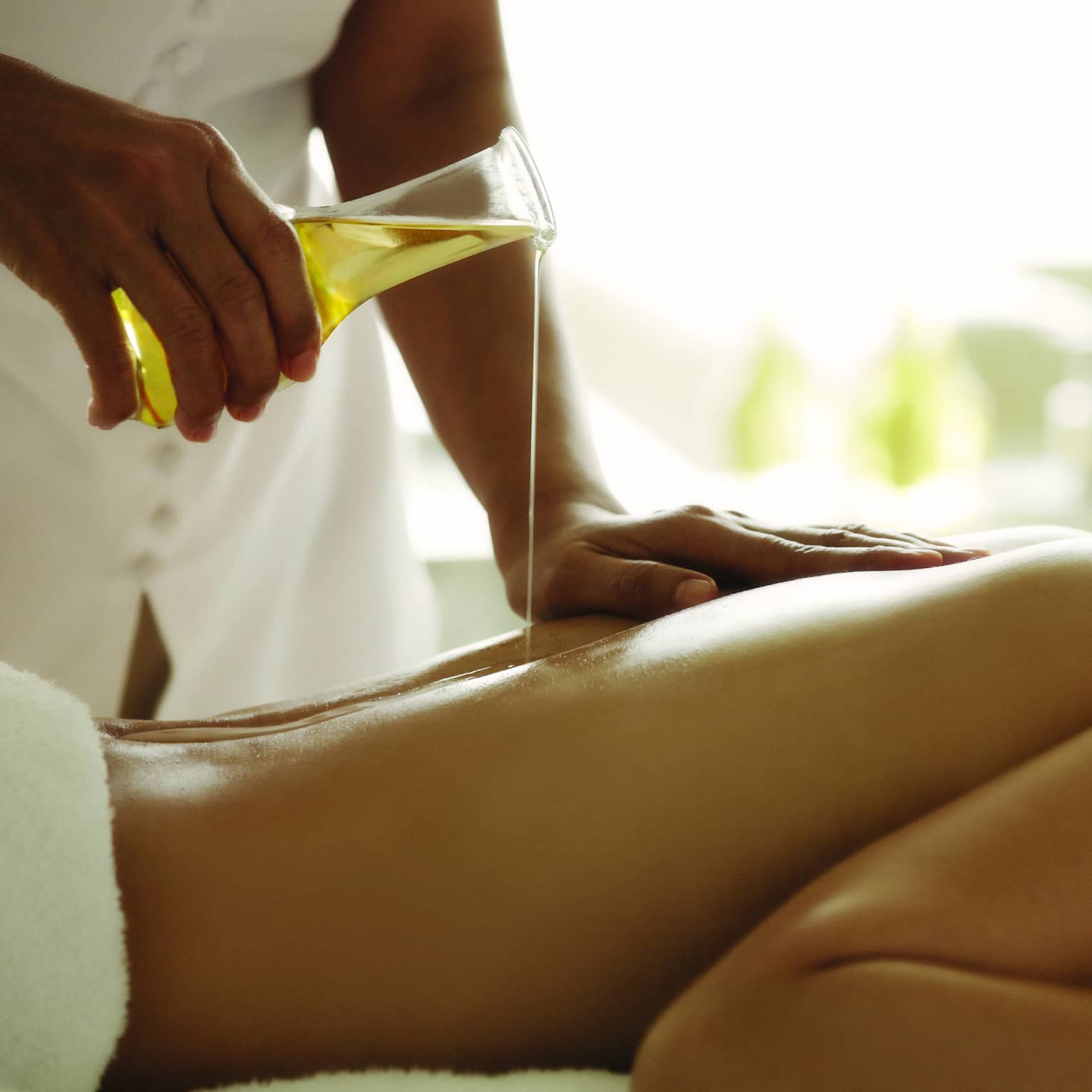 Spa staff pours massage oil over woman's bare back as she lays on treatment bed