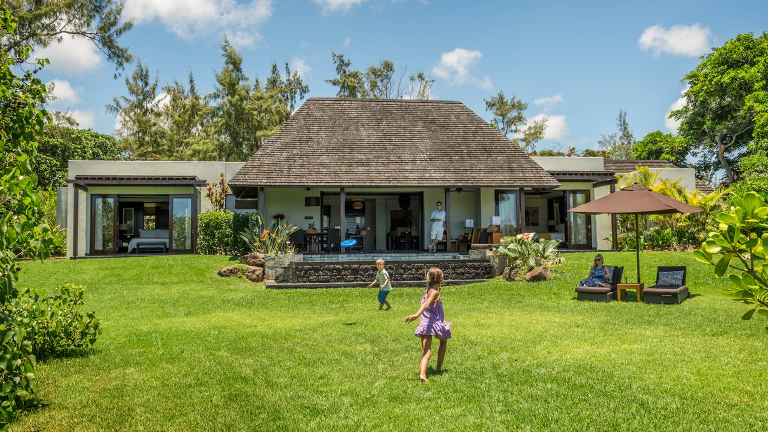 Young children play on green lawn in front of bungalow as dad watches from covered patio