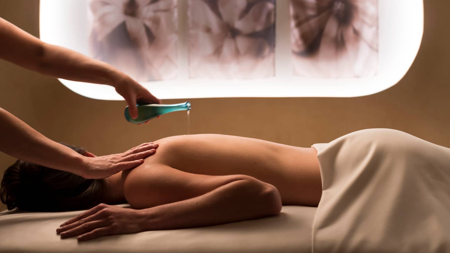Woman's hands holding blue bottle pours liquid on woman's bare back in spa room