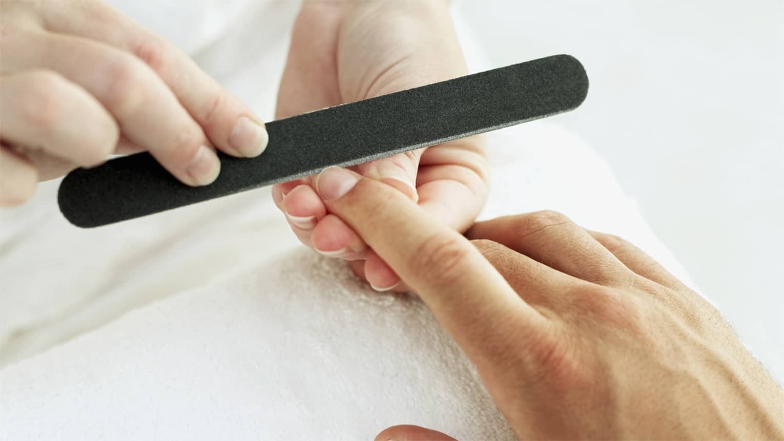 Woman holds manicure nail file to man's fingernails over white towel
