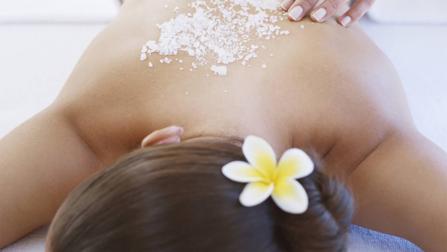 Woman lying on massage table with white jasmine flower in hair, salt scrub on bare shoulders