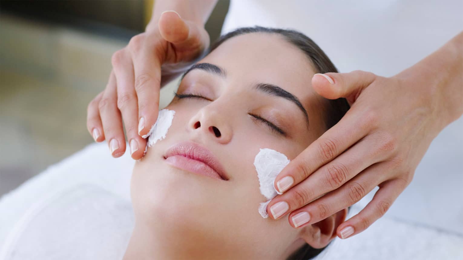 Woman closes eyes as spa staff rubs cream on cheeks