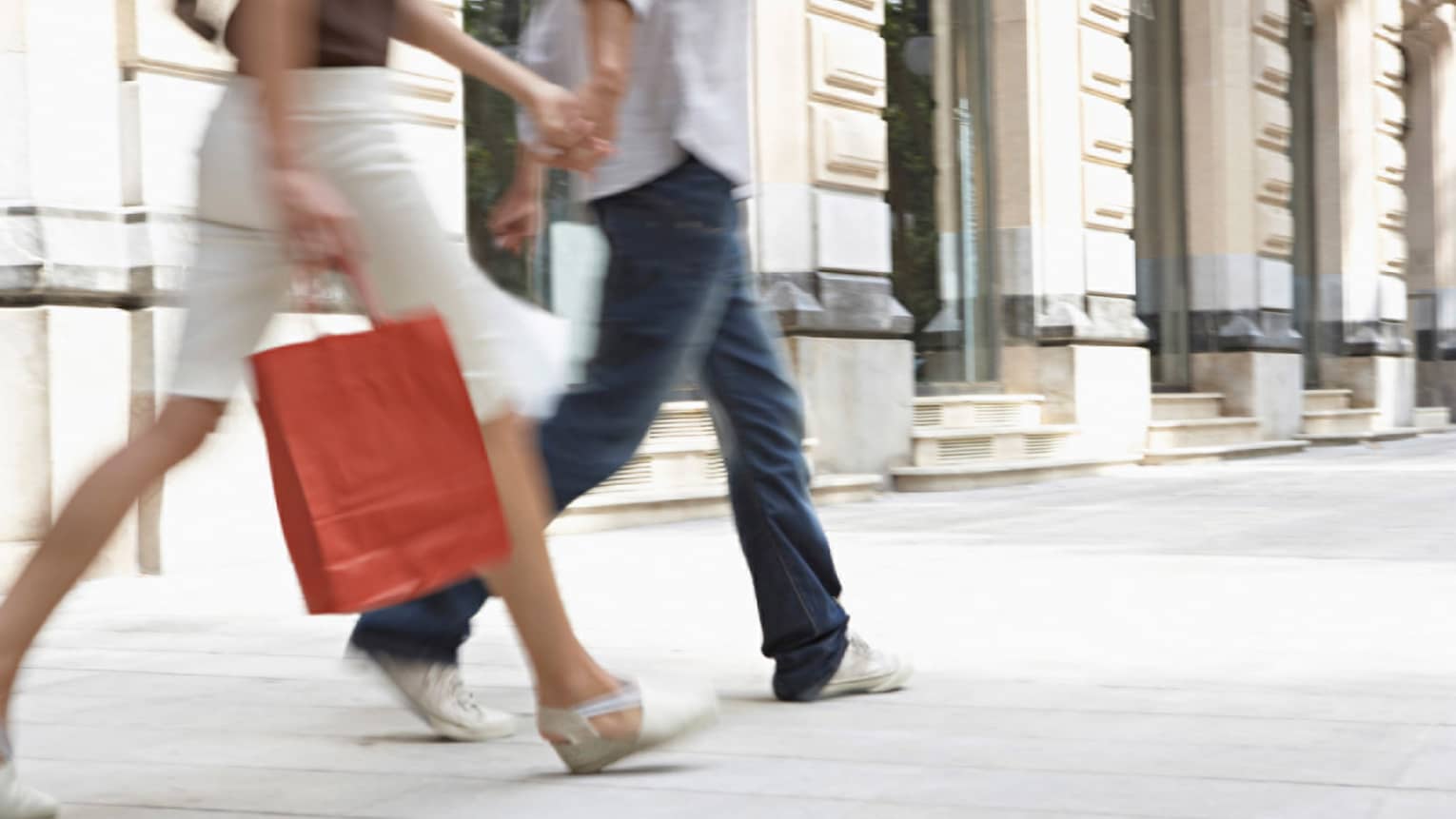 Out-of-focus lower half of couple holding shopping bags, walking down city sidewalk in summer