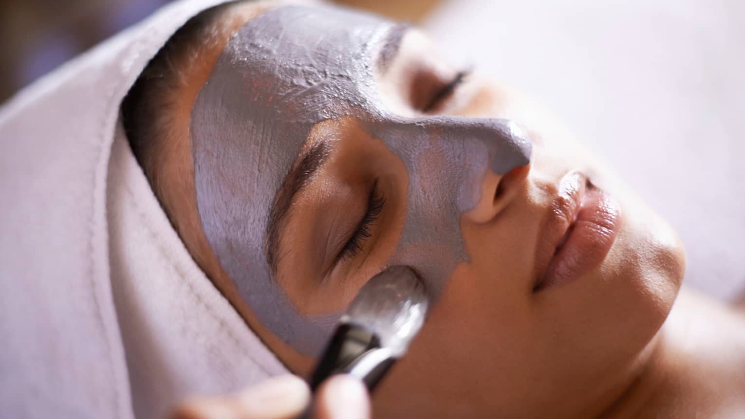 Woman with white towel around hair closes eyes as spa attendant applies grey face mask with paintbrush