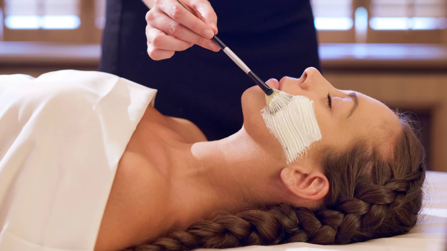 Woman with eyes closed lies on back on spa table as woman with paintbrush applies Sodashi-based facial cream