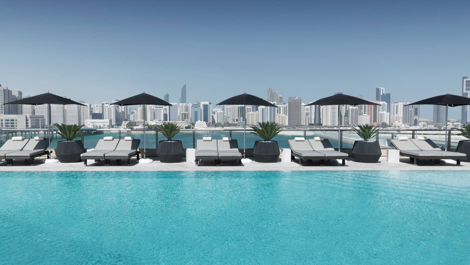 A row of light-grey lounge chairs lines an outdoor pool with skyline views in the background