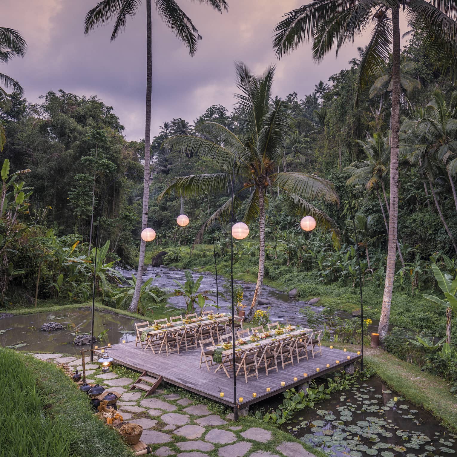 Two long tables laid out with food sit on a lantern-lit platform above a lotus pond and beside a river and dense forest.