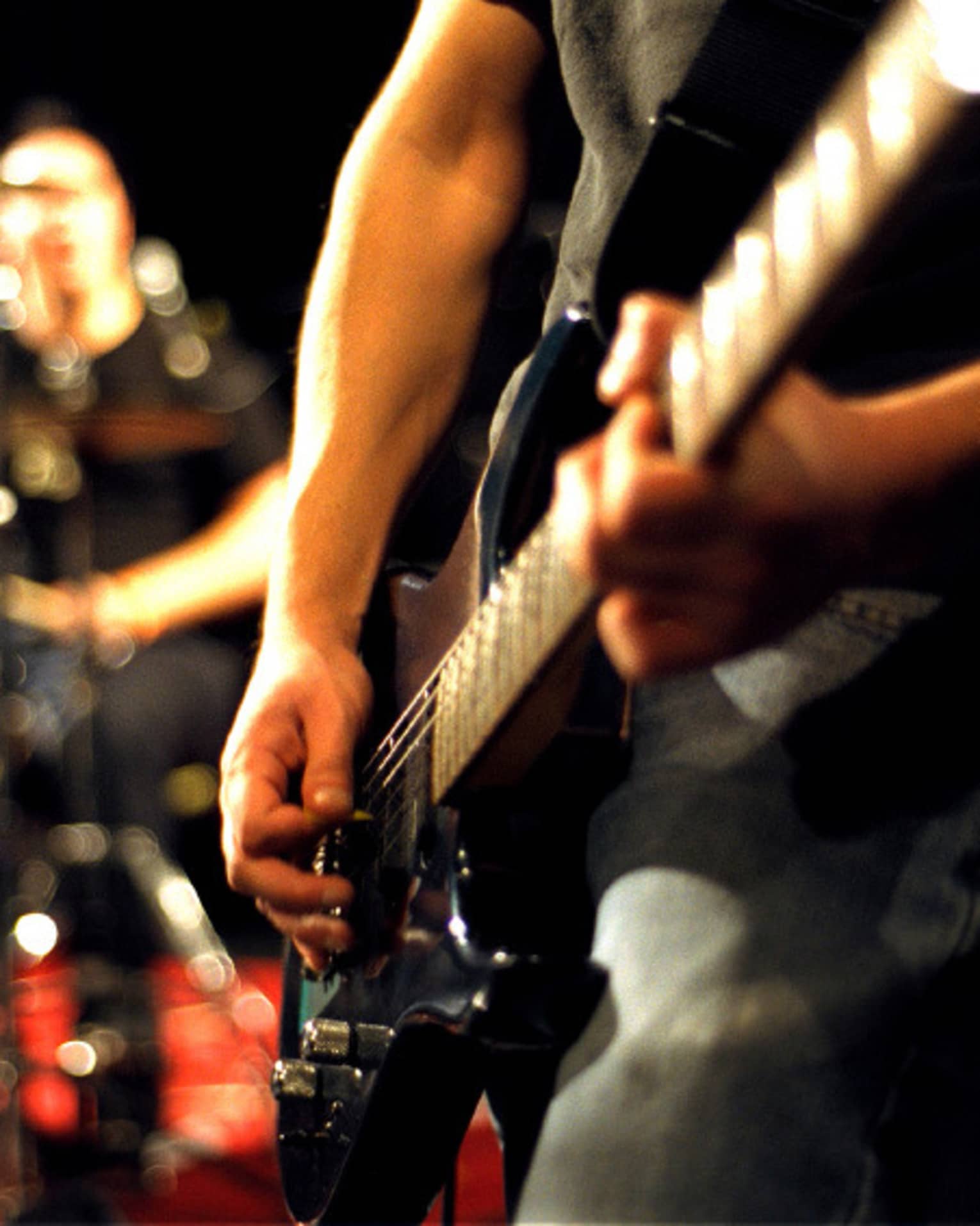 Close-up of musician's arms and hands playing an electric guitar with drumset in the background.