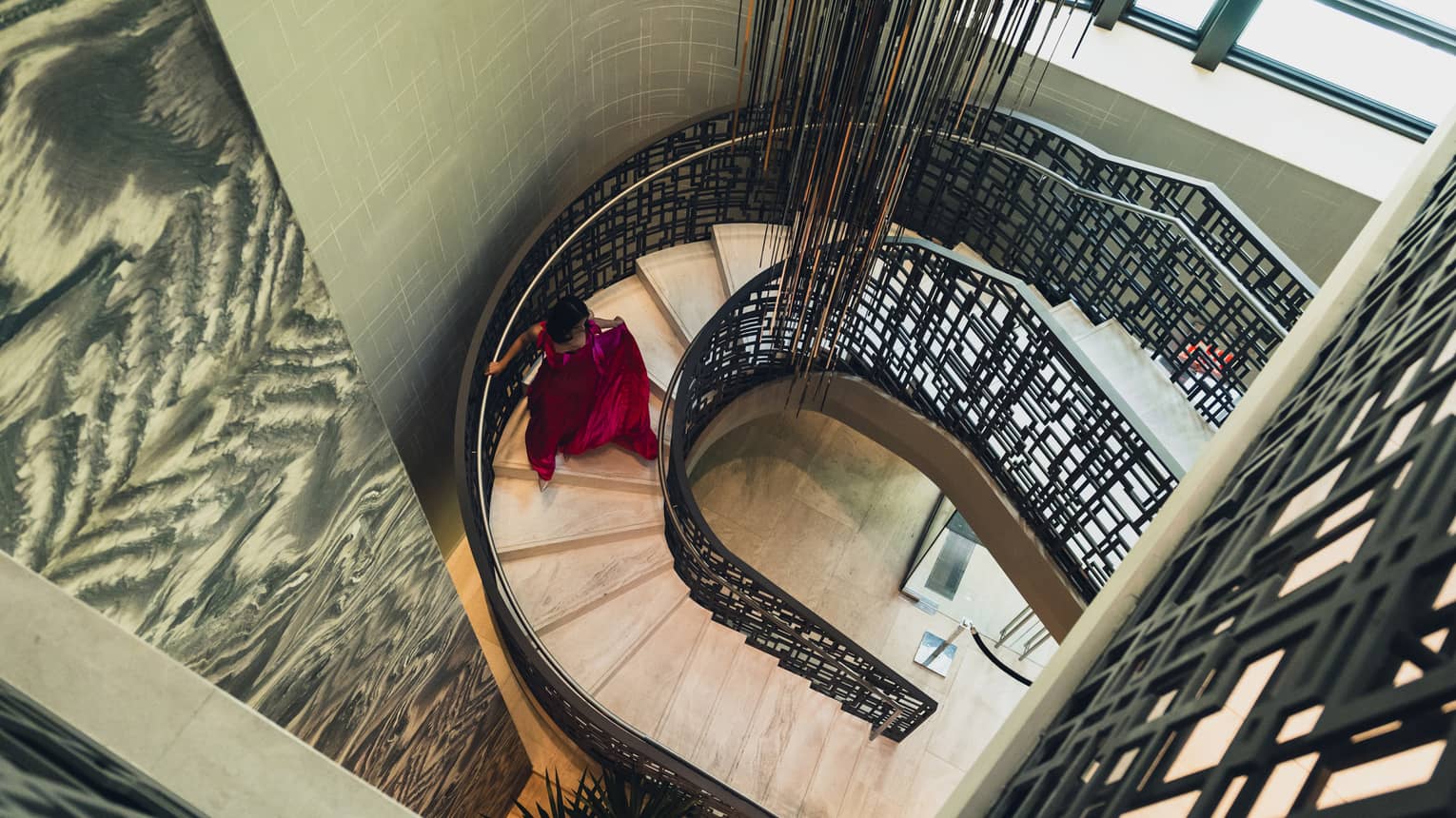 Woman in red evening gown descends a spiral staircase, light streaming in from the window