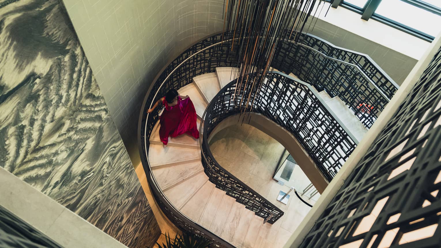 Woman in red evening gown descends a spiral staircase, light streaming in from the window