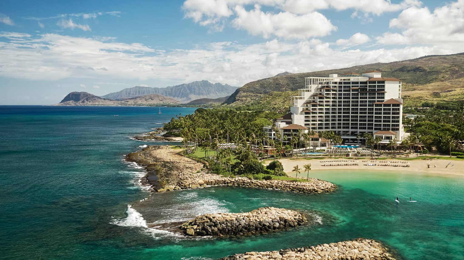 Tall white Four Seasons Resort Oahu building on mountainside by sand beach, rocks, ocean