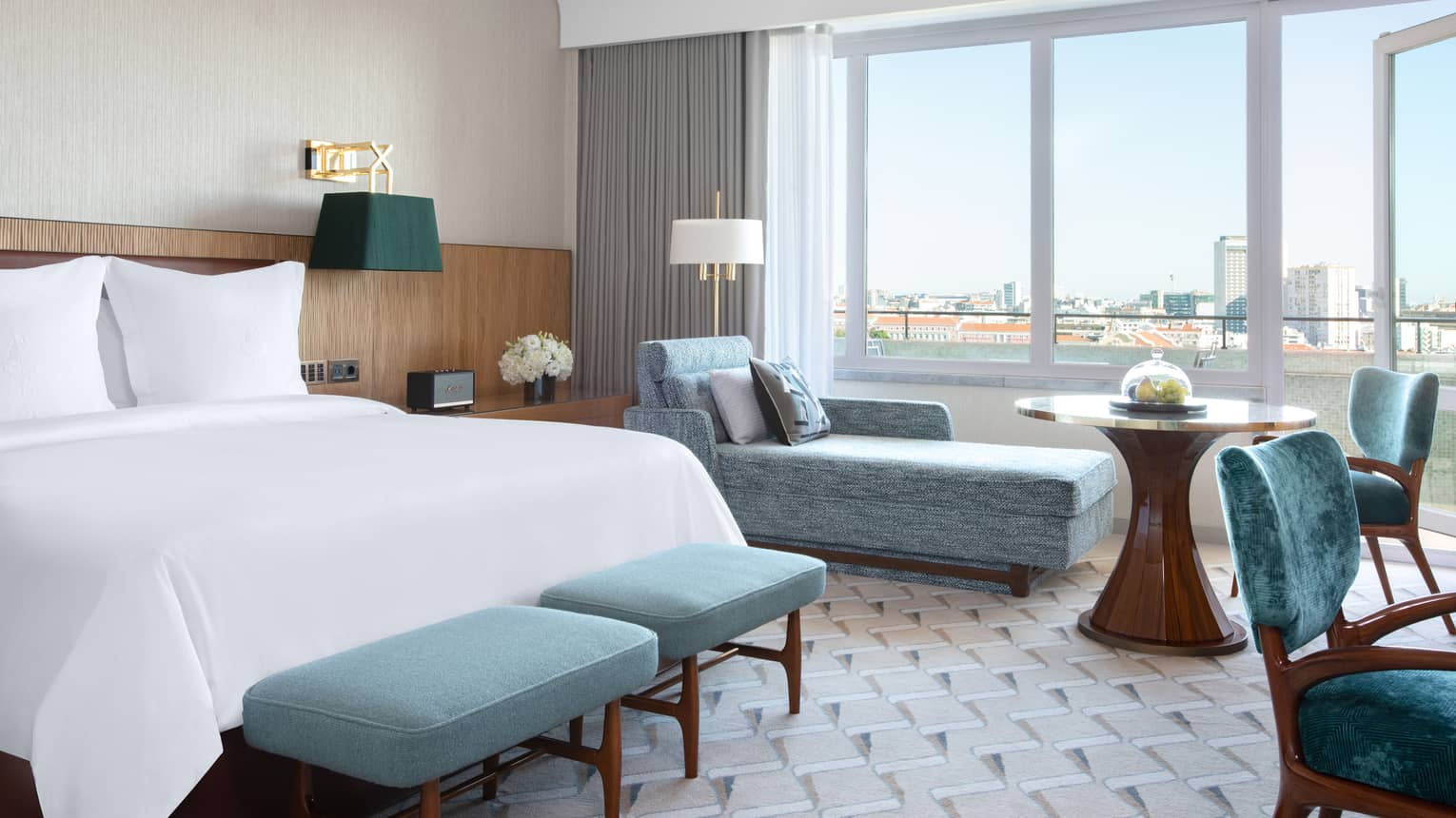 Guest room with white bed, blue benches at foot, blue chaise longue, tall windows with view
