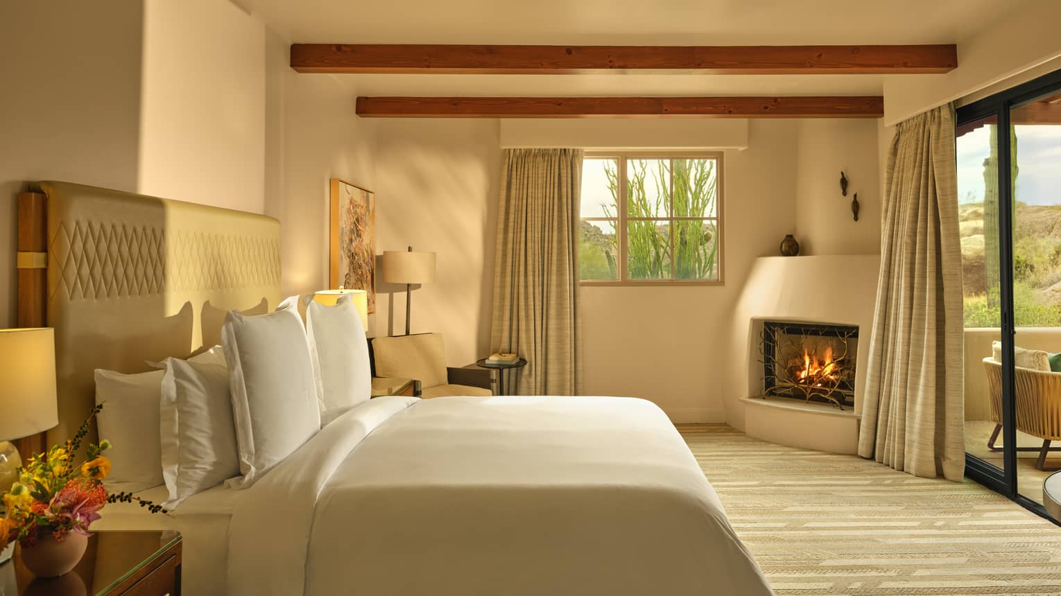 Light streams into a bedroom of luxury suite, with a king bed, fireplace, beamed ceiling and walk-out terrace