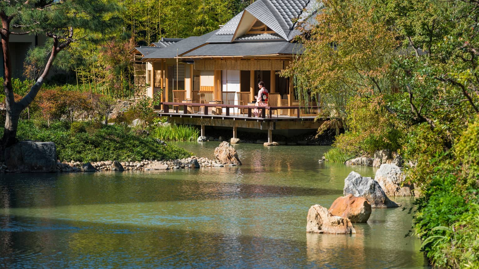 View across pond to bungalow where woman in robe sits on patio