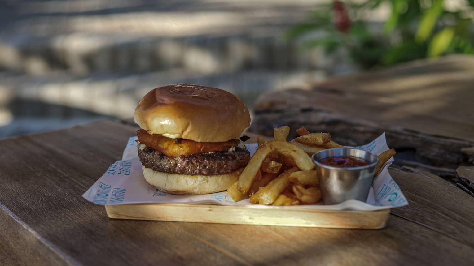 A small rectangular wooden platter holds a hamburger in a bun, French fries and a small steel container of ketchup on a wooden picnic table