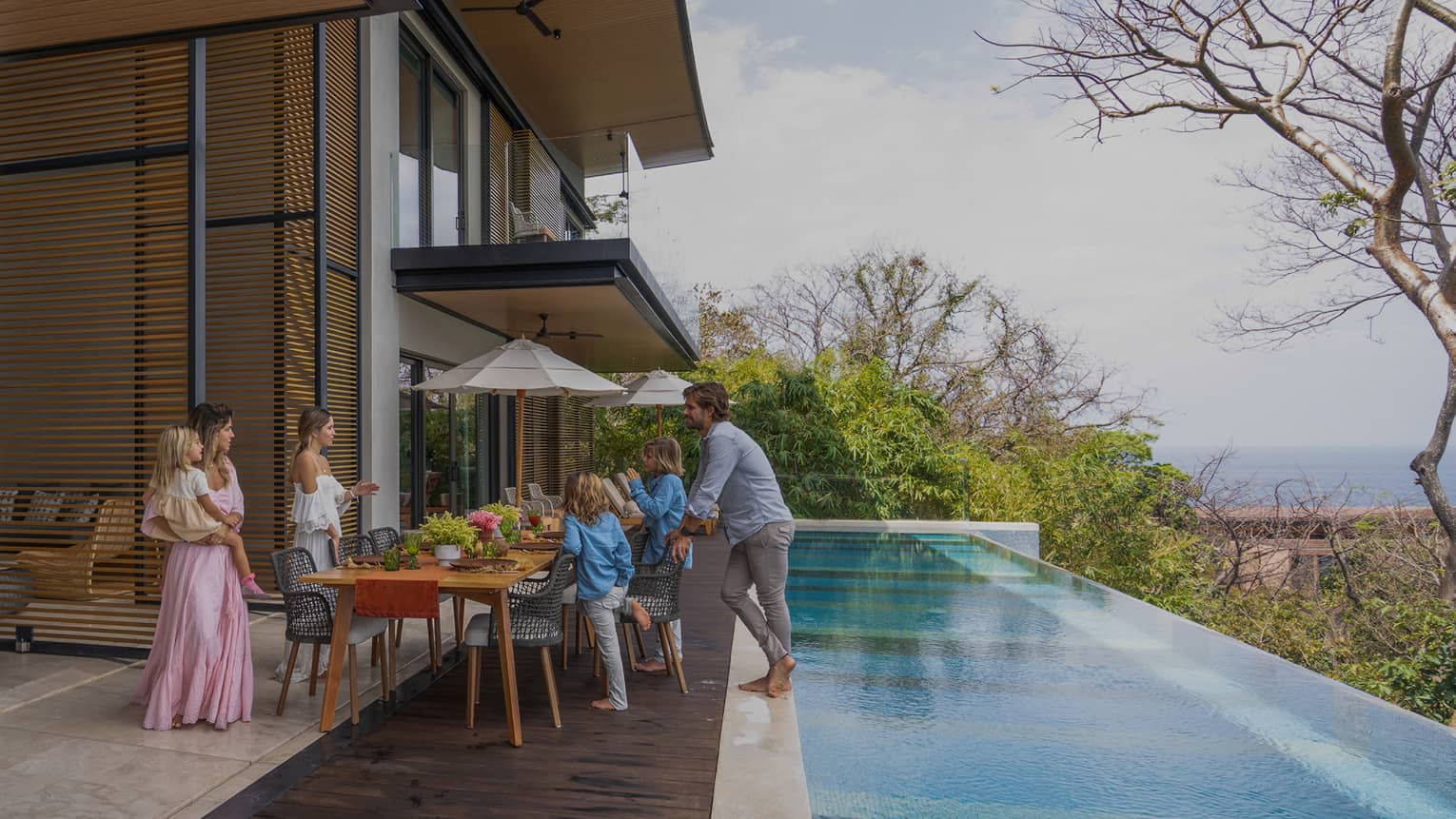 Family gathers around dining table set outside on a deck next to a private infinity-edge pool