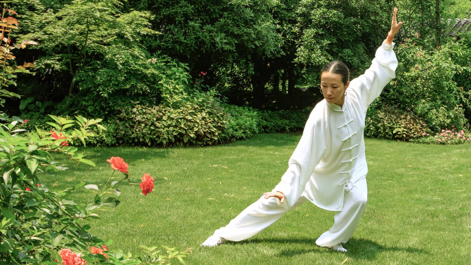 An instructor in a white tai chi outfit, arms and legs outstretched in a classic pose, in front of a pagoda-shaped gazebo.