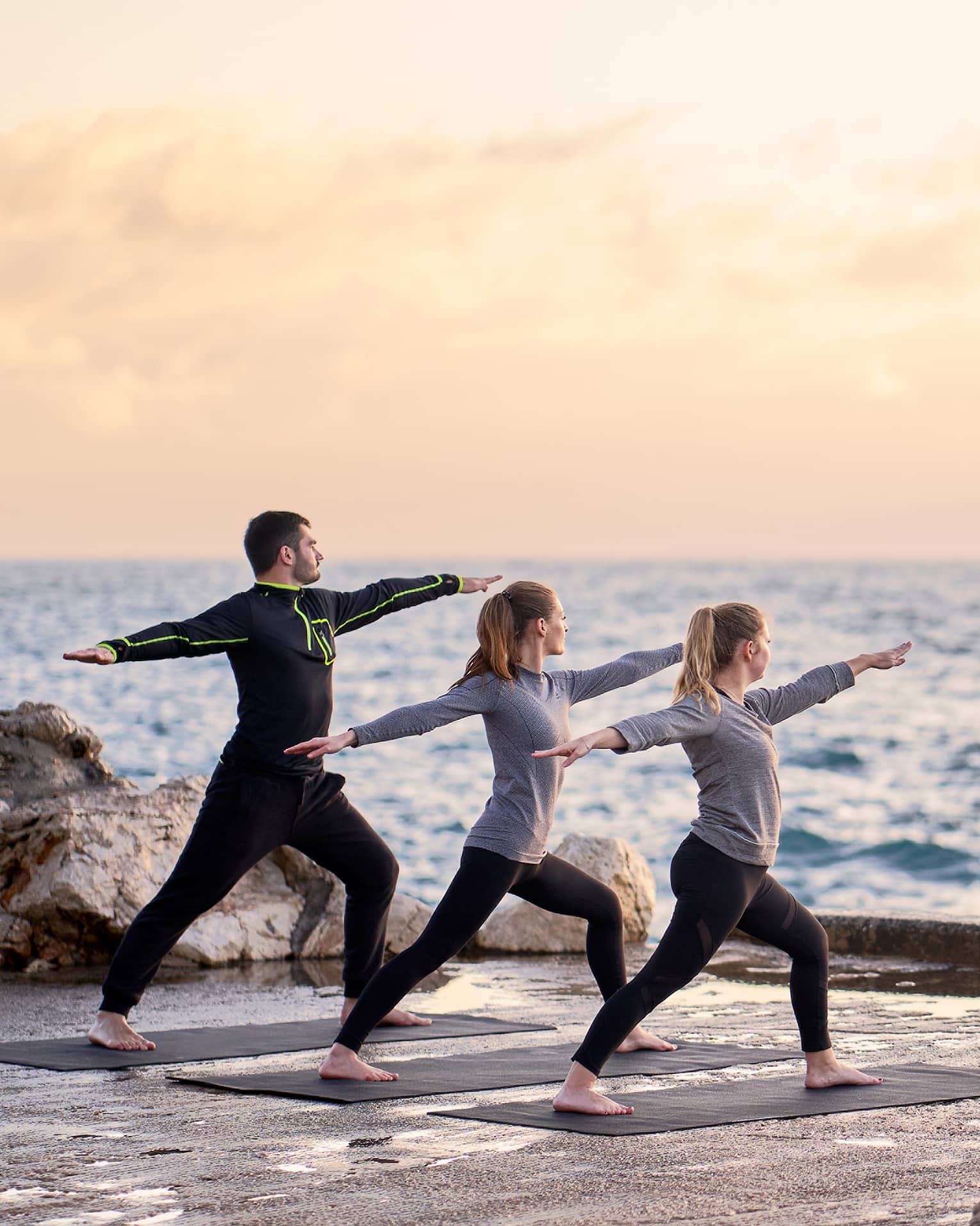 Three people in warrior yoga pose and coach standing at sea's edge at sunrise