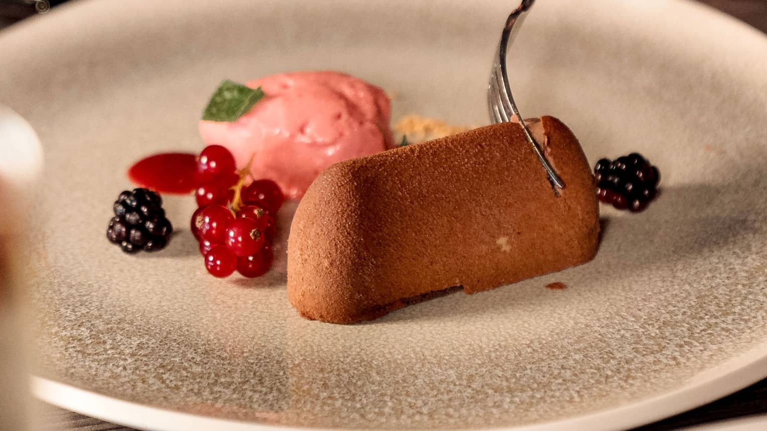 A person slides a fork into a pastry garnished with fruit and served with a pink sorbet