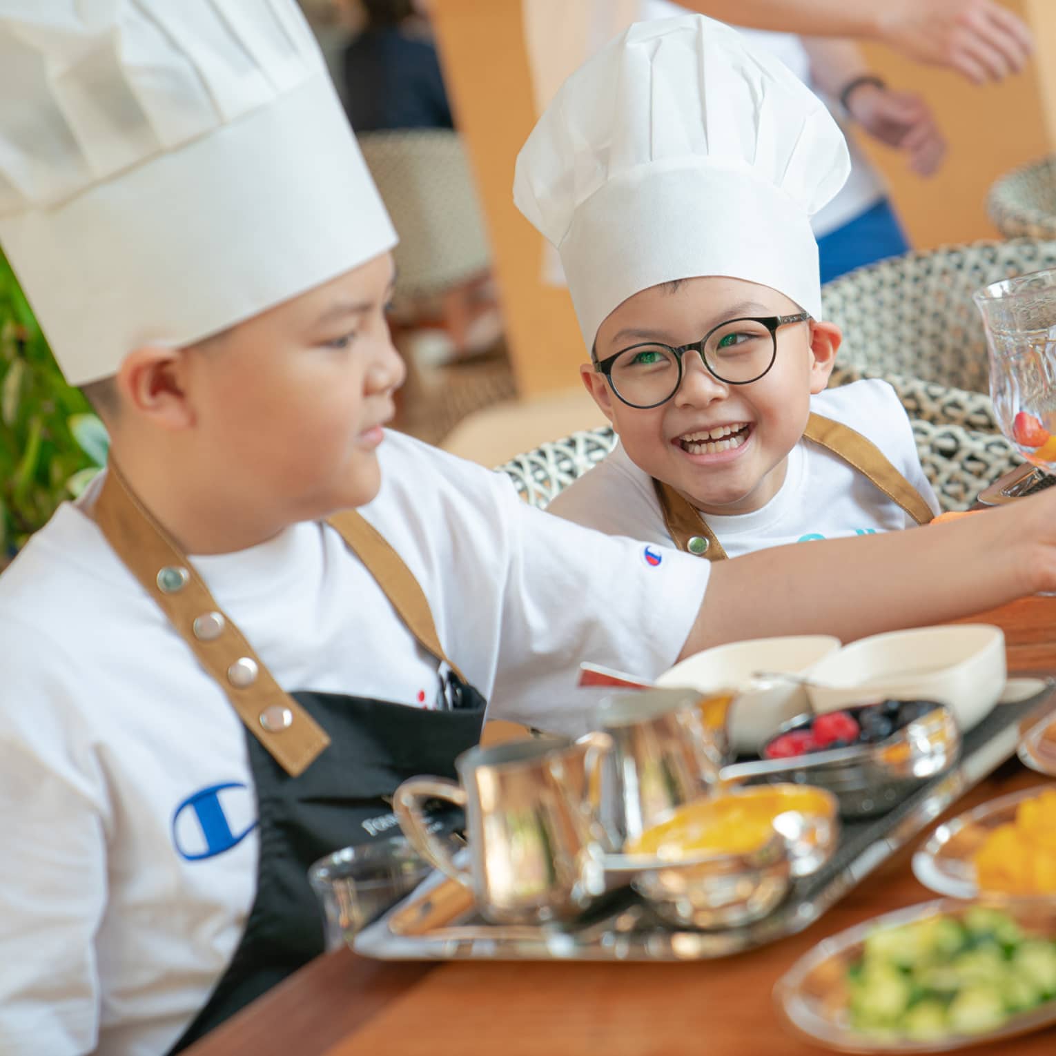 Two children wearing white chef's hats, white t-shirts and blue aprons sit at a table in front of various small plates of ingredients