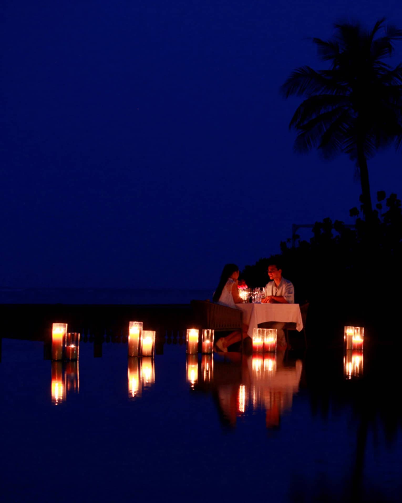 Couple dine by lagoon at small table surrounded by glowing candles at night