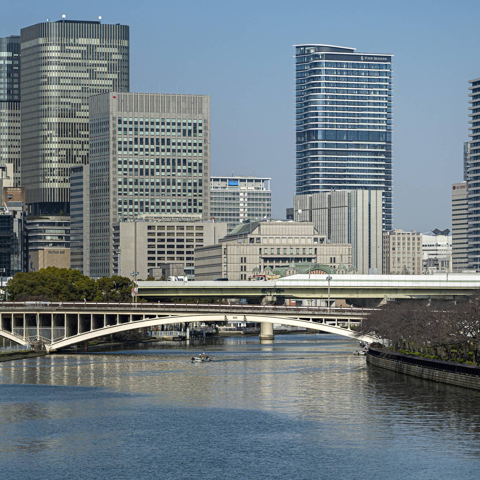 Under a clear blue sky, Osaka's modern high-rises loom above two bridges spanning a river lined with dormant cherry blossoms.