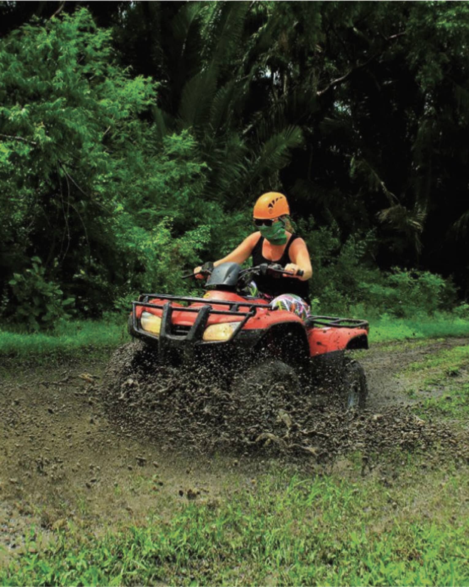 Person wearing a black tank top and bright orange helmet rides through the mud on a red ATV