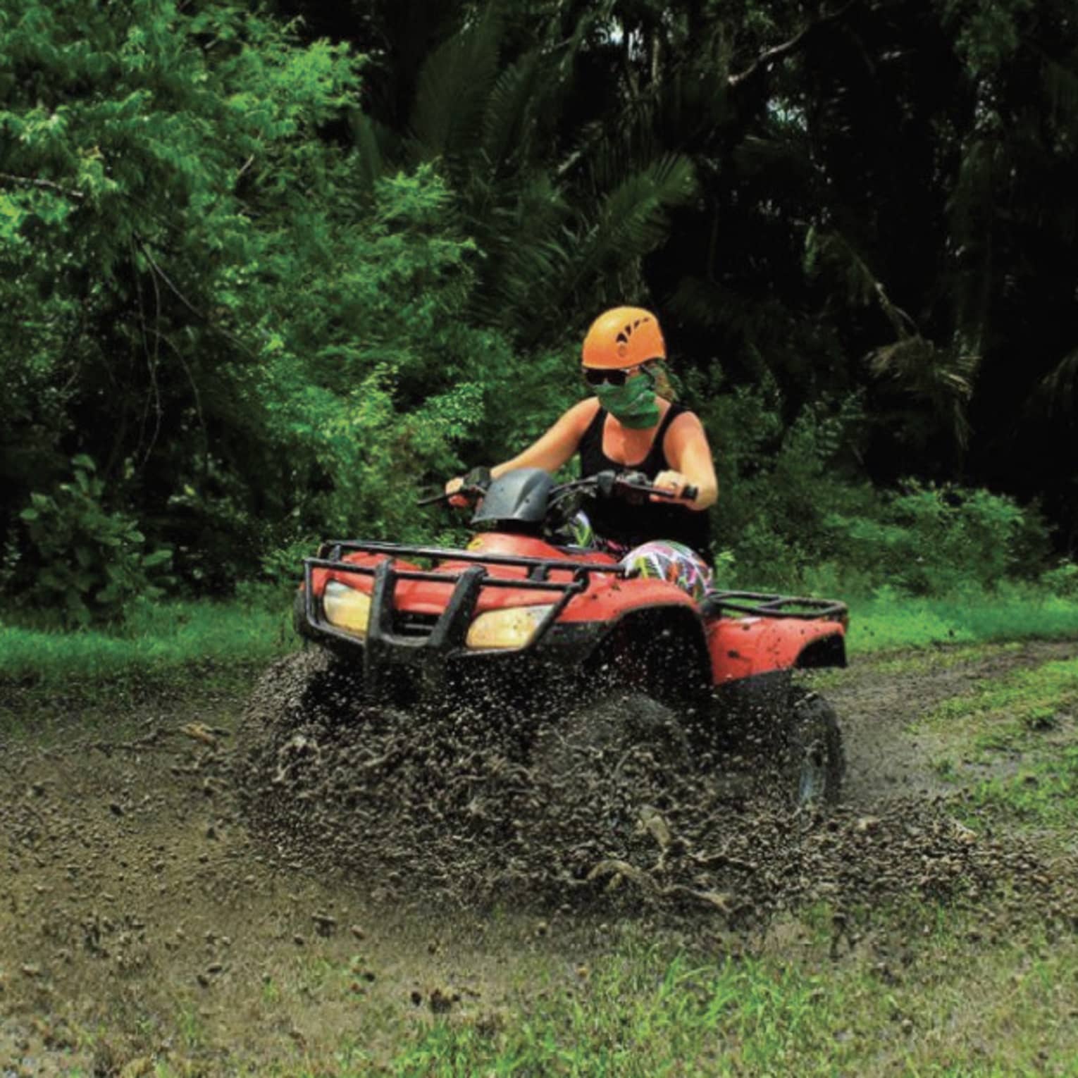 Person wearing a black tank top and bright orange helmet rides through the mud on a red ATV