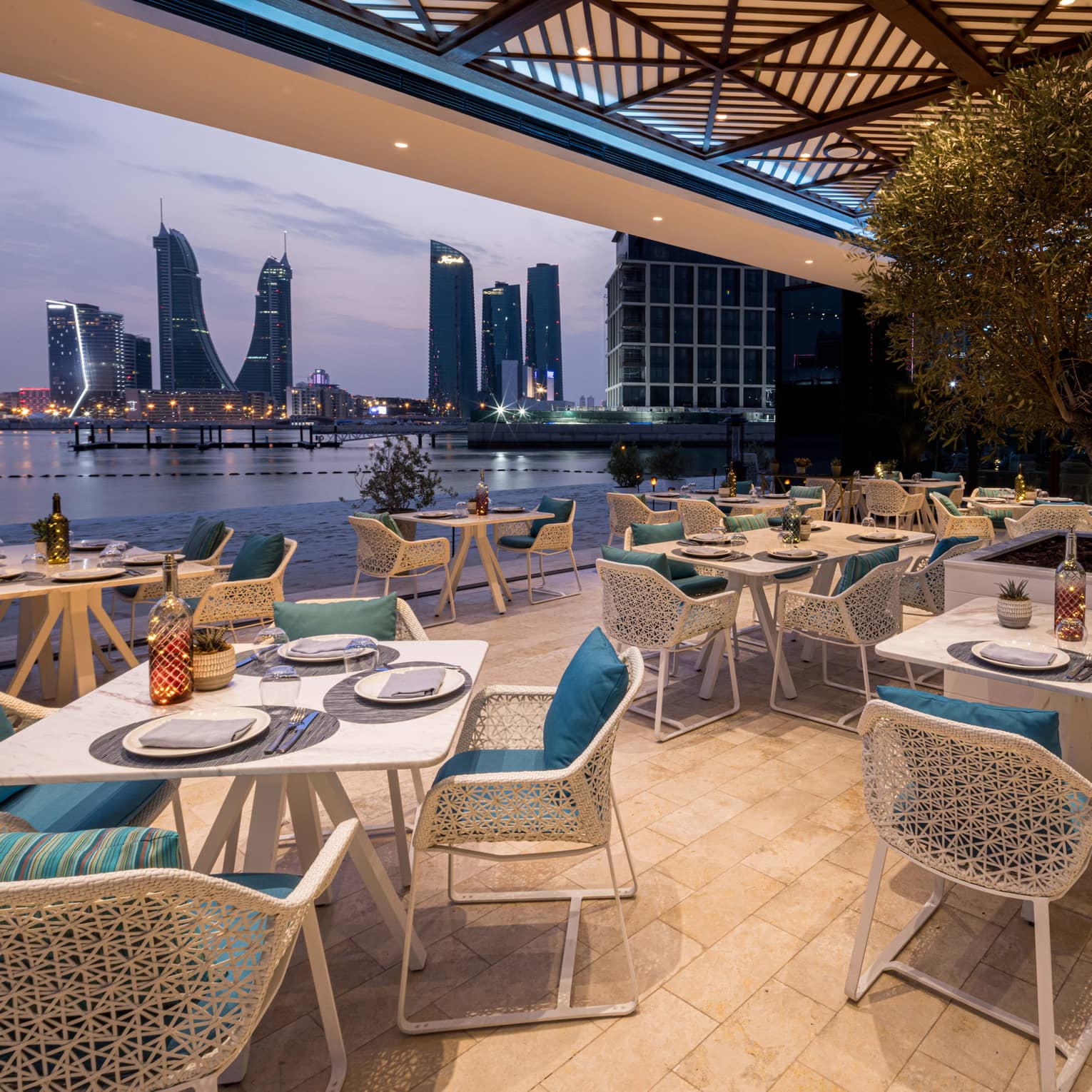 Restaurant terrace with white tables and chairs with blue cushions opens up to view of the water and a city skyline