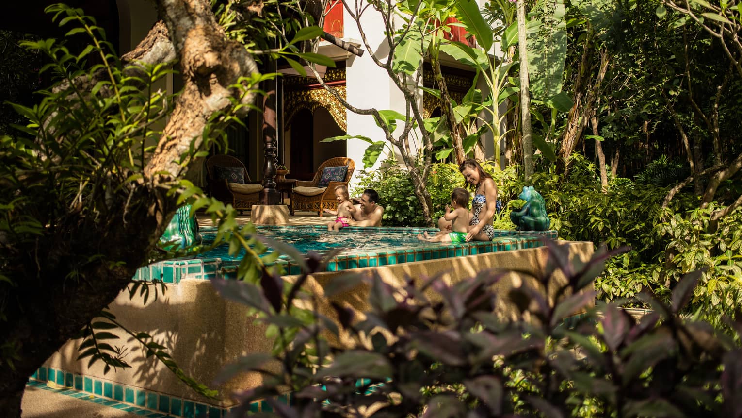 Couple with two babies sit on edge of private pool in front of bungalow by garden