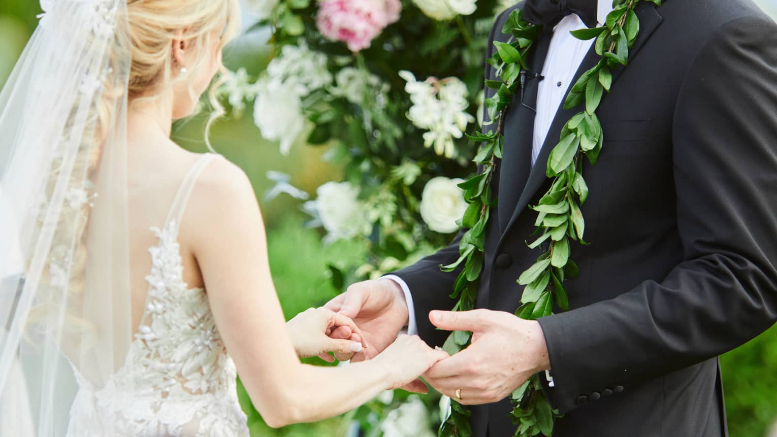 A couple saying their vows beneath a large floral arch. The woman has a chapel length veil and a lacy dress, the man is wearing a black suit with a floral garland around his neck.