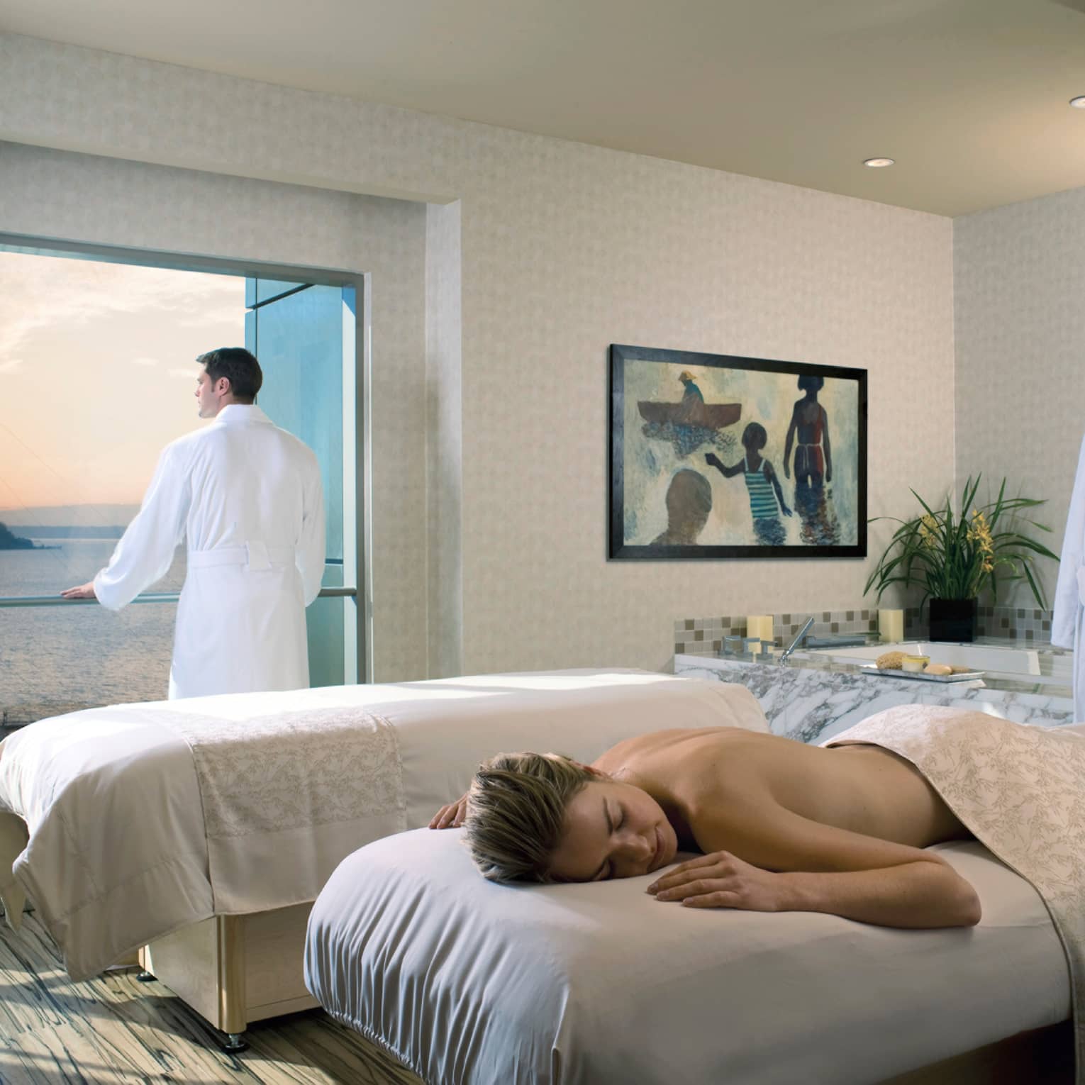A couple's spa treatment room, woman laying on massage table, man wearing bathrobe looking out window